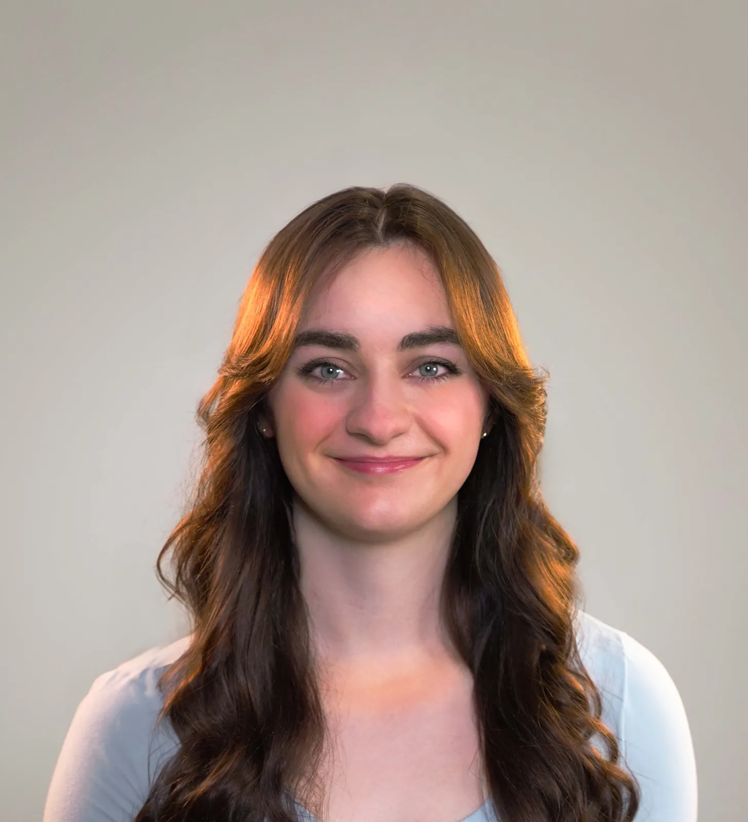 Close-up, well-lit smiling young woman with long brown hair in a light neutral studio setting.
