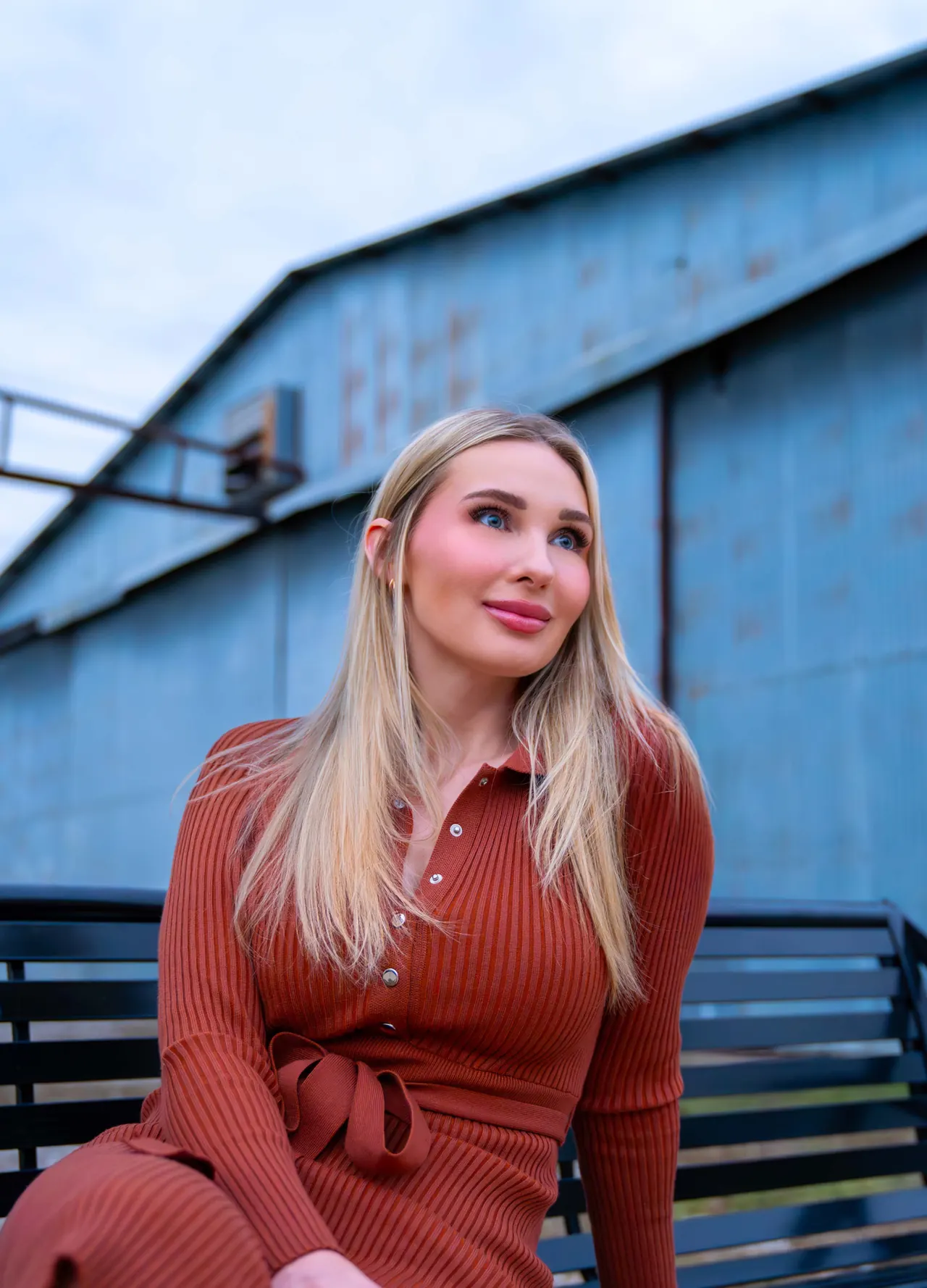 Portrait of a woman in a rust knit dress seated in front of a blue metal barn, by Dark Rainbow Photo Video